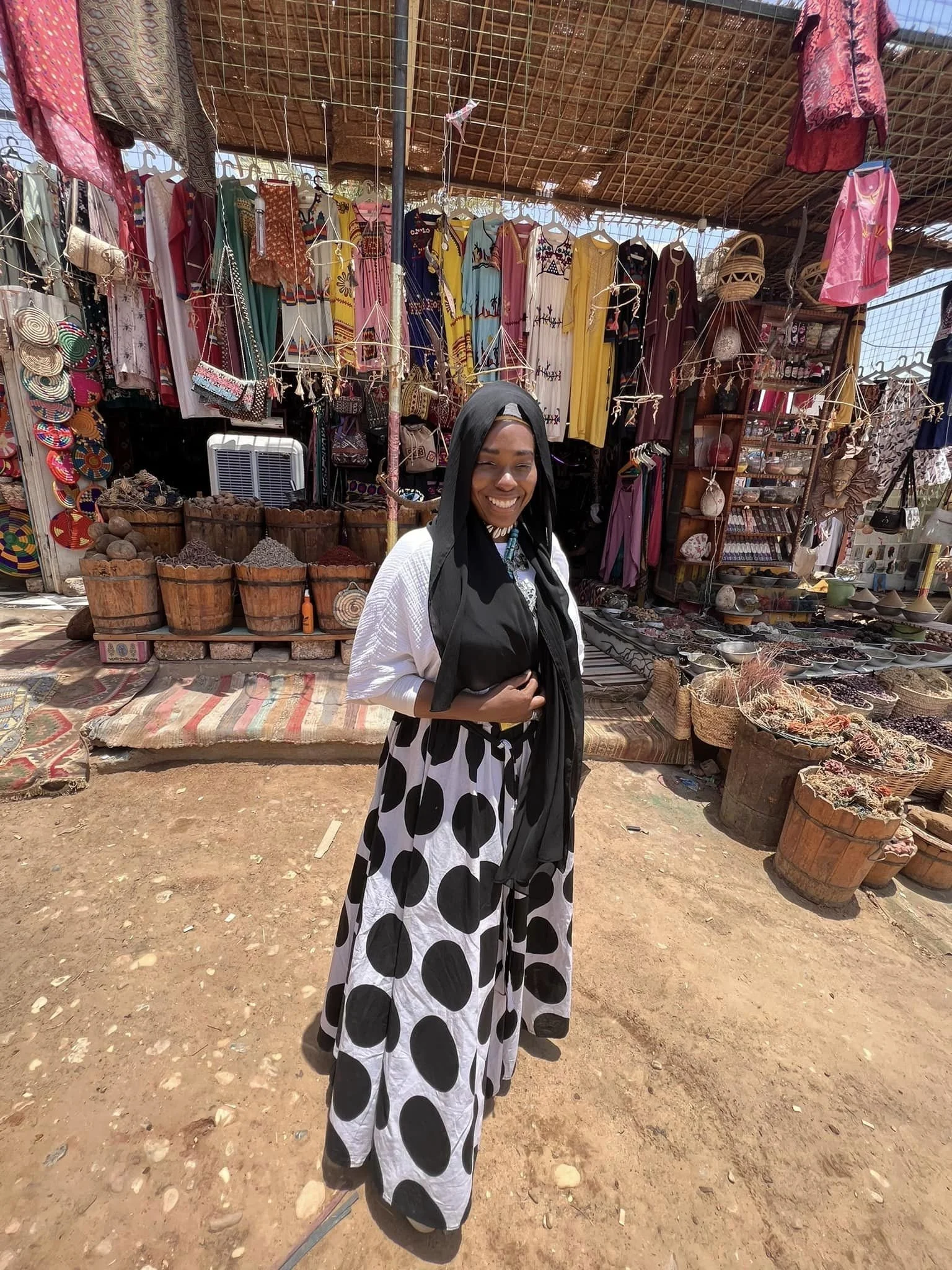 A woman wearing a black headscarf, white blouse, and long black and white polka dot skirt stands smiling at an outdoor market with colorful garments, textiles, and pottery in the background.