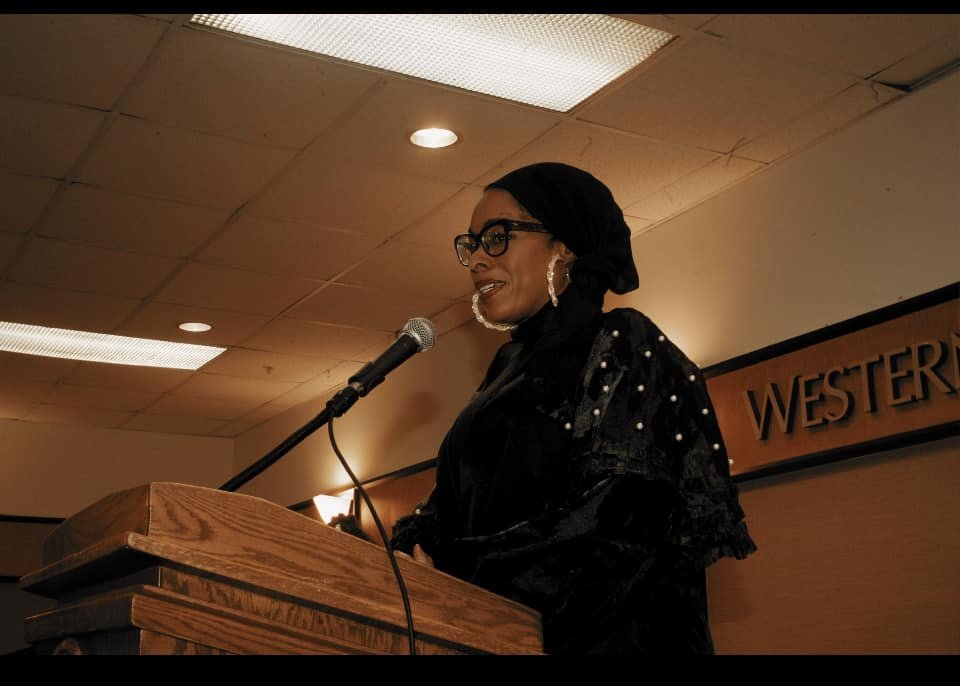 A woman wearing glasses and large earrings is speaking at a podium with a microphone inside a room with ceiling lights and a sign that reads Western.