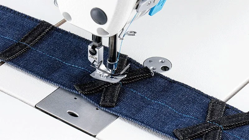 Close-up of a sewing machine stitching denim fabric with a blue seam, creating a belt loop.