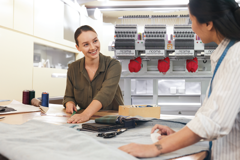 two women in an embroidery shop smiling with a Ricoma 4-head embroidery machine in the background