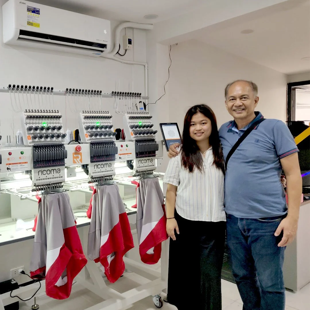 A young woman and an older man smiling and standing next to each other indoors, with embroidery machines and a wall-mounted air conditioner in the background.