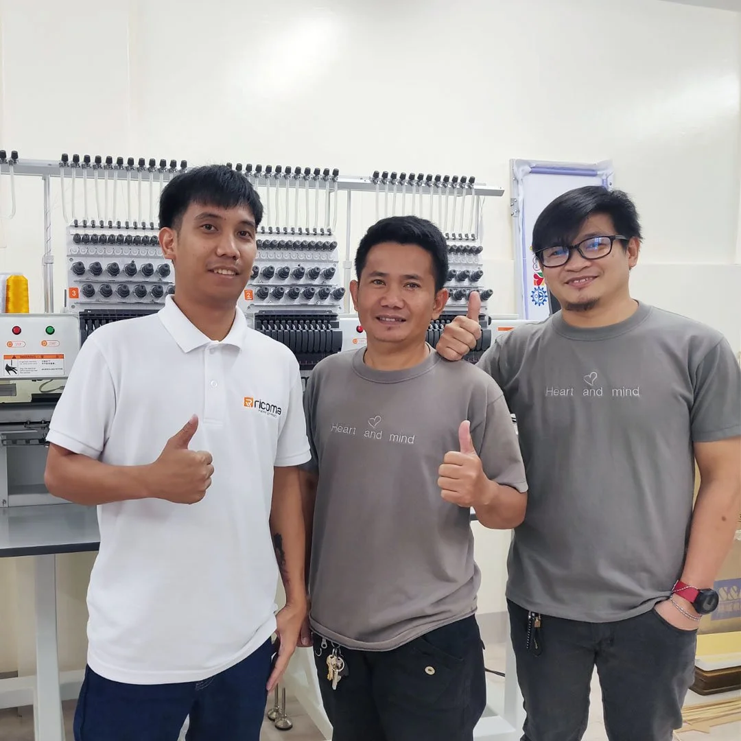 Three men standing indoors, smiling and giving thumbs up, behind embroidery or sewing machine equipment.