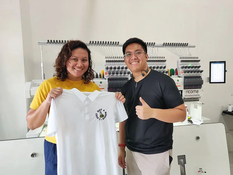 Two smiling people in a textile workshop holding a white polo shirt with a logo that reads "Just Do It" and a graphic of a hand giving a thumbs-up symbol. There is a Ricoma 4-head embroidery equipment in the background.