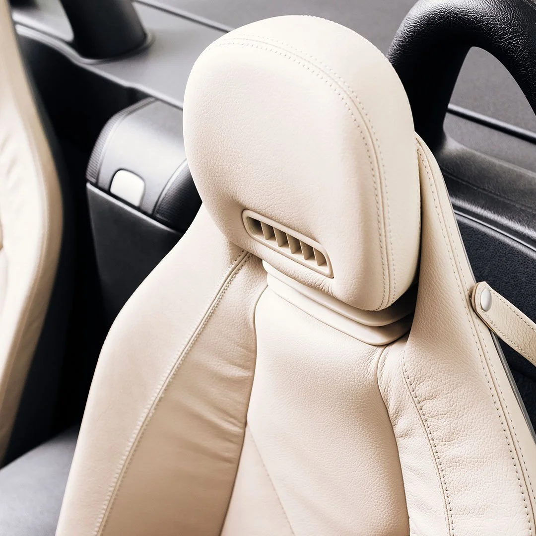 Close-up of a beige leather car seat headrest with an adjustable vent or air circulation vent behind it, and part of a black steering wheel in the background.