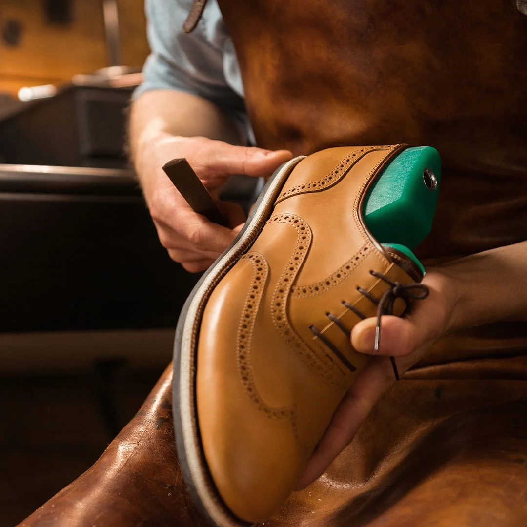 A person is polishing a tan leather dress shoe with brogue detailing, using a green polishing tool inside the shoe.