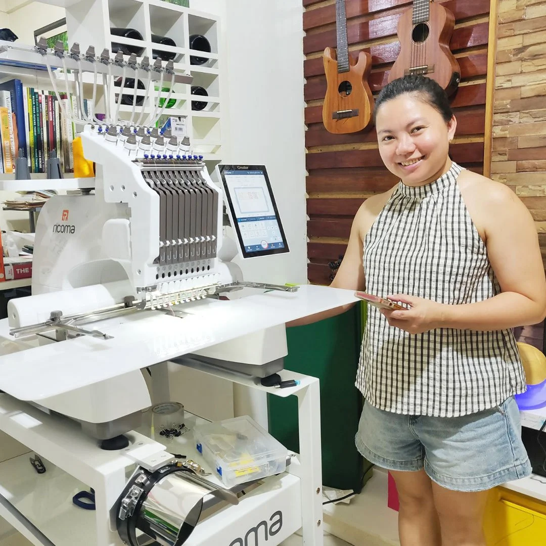 A woman in a checkered sleeveless top and denim shorts standing next to an embroidery machine in a room with musical instruments hanging on the wall.
