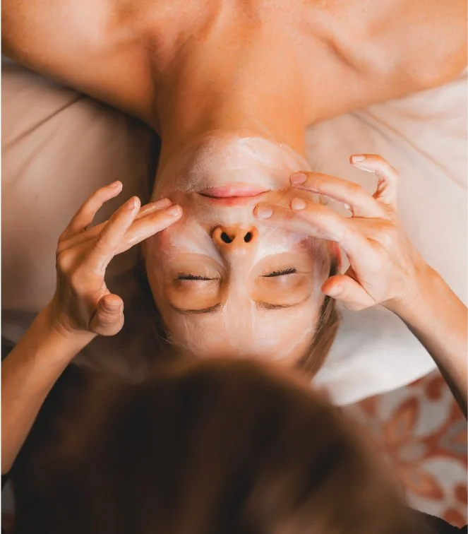 Person lying down with eyes closed, receiving a facial treatment, with a white facial mask applied and hands gently touching the face.