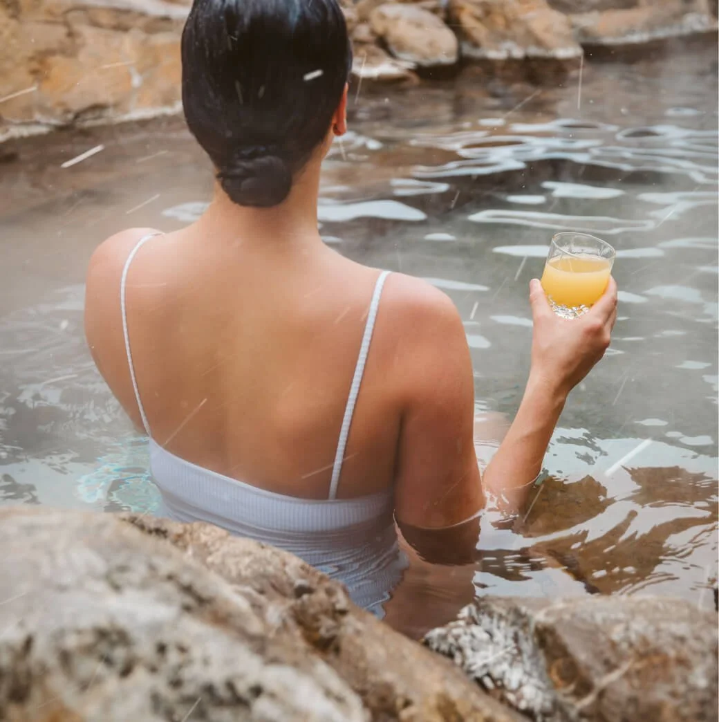 A woman with dark hair tied back in a bun is sitting in a hot spring or natural pool, holding a glass of orange juice. She is wearing a white swimsuit with thin straps and is surrounded by rocks with water dripping down them.