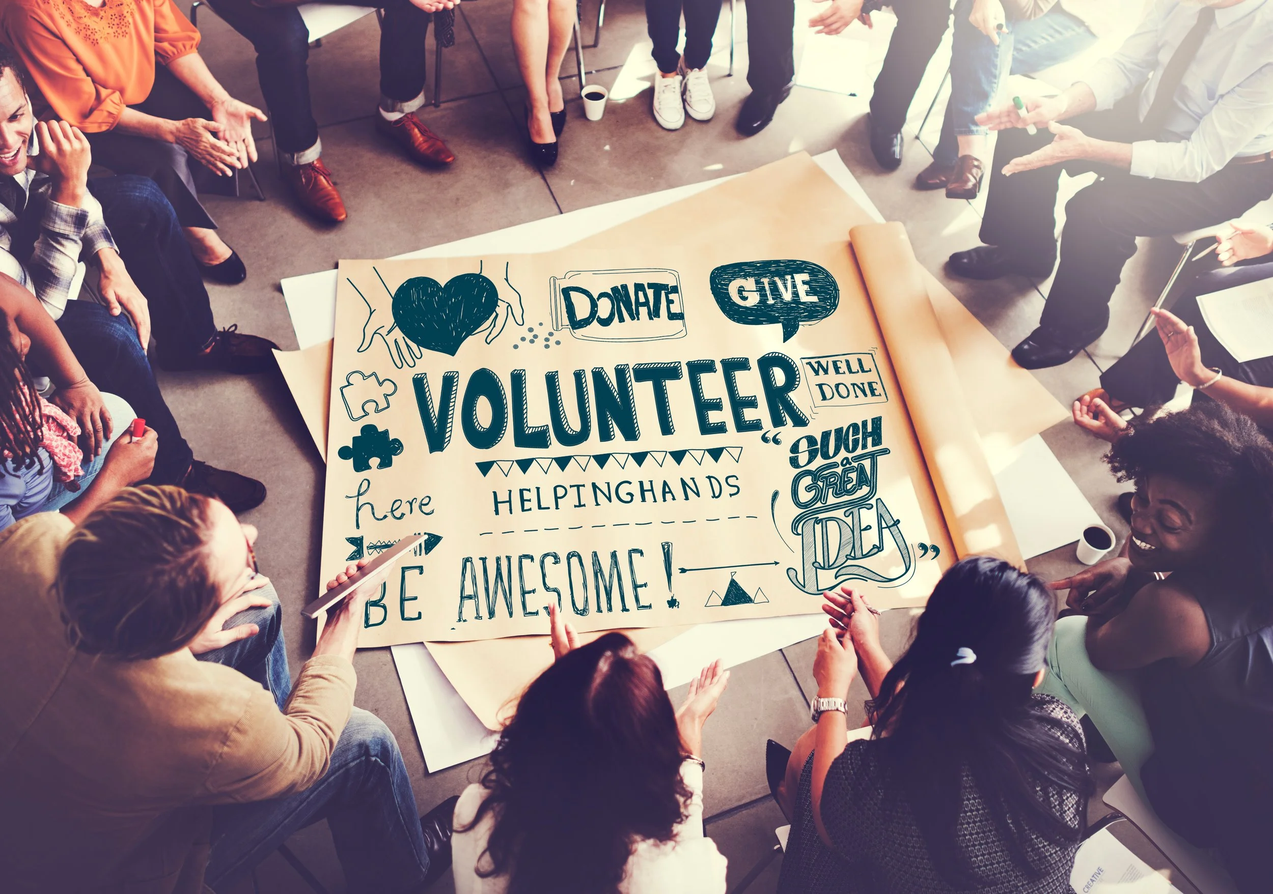 A group of diverse people sitting in a circle around a large poster with handwritten messages about volunteering, fundraising, and helping hands, in a community or meeting room.