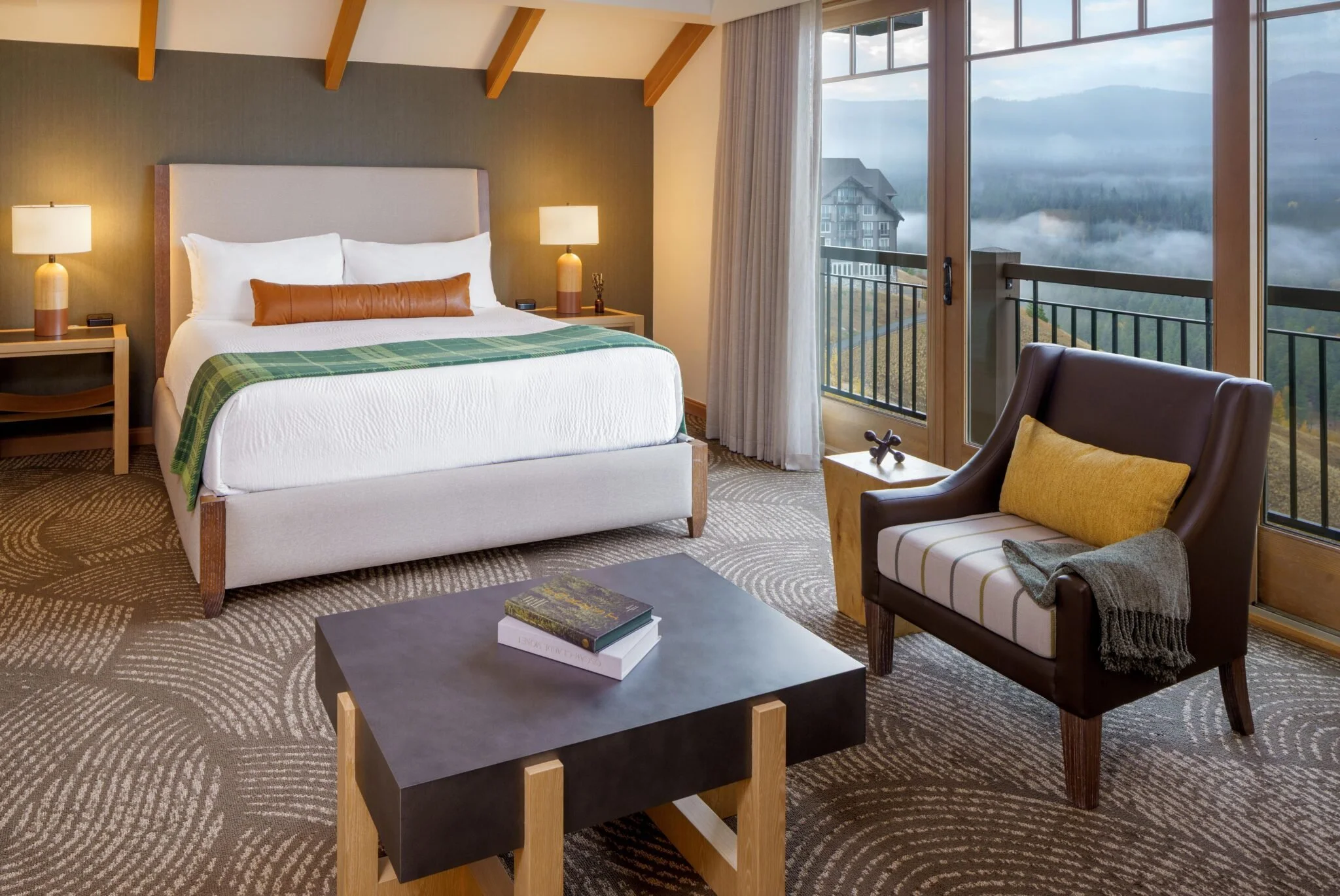 A cozy hotel room with a large bed, bedside tables with lamps, a sitting area with an armchair and blanket, a coffee table with books, and a sliding glass door leading to a balcony with scenic mountain views.