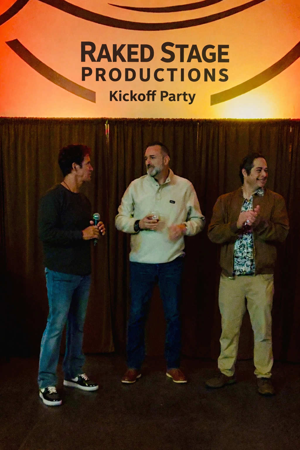 Three men are standing on a stage at a kickoff party for Raked Stage Productions. The man on the left is holding a microphone, the man in the middle is holding a drink, and the man on the right is clapping. A large sign with the company's name and event title is displayed above them.