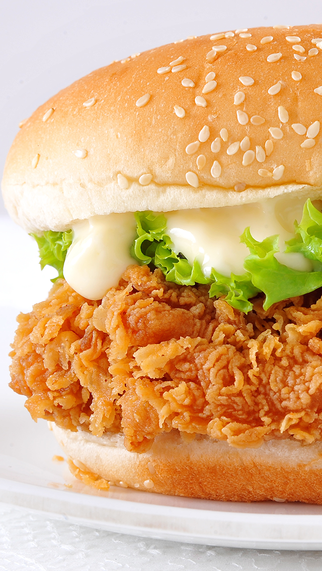 Close-up of a fried chicken sandwich with lettuce, mayonnaise, and a sesame seed bun on a white plate.