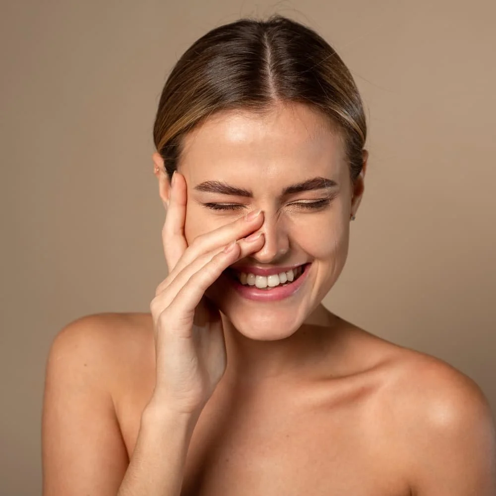 A woman smiling and touching her nose with her hand, with a neutral background.