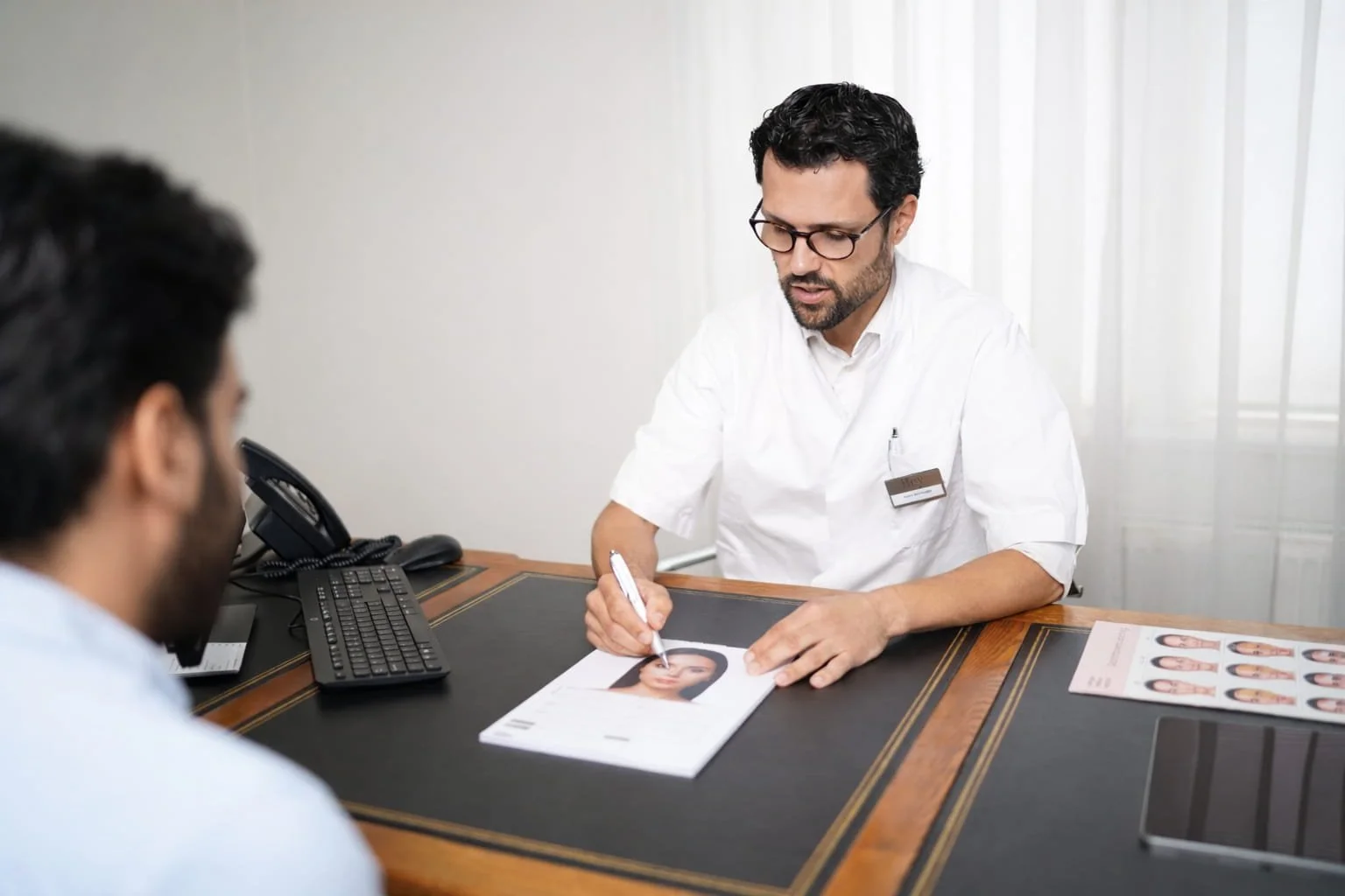 A man with glasses and a beard sitting at a desk with a photo ID, talking to a person whose back is to the camera. The desk has a computer, a telephone, and photo sheets, with light-colored curtains in the background.