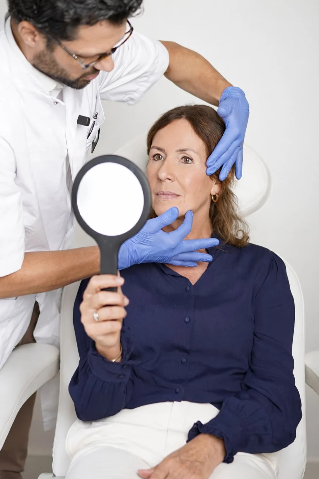 A female patient sitting upright, holding a mirror, as a doctor examines her face with gloved hands in a medical office setting.