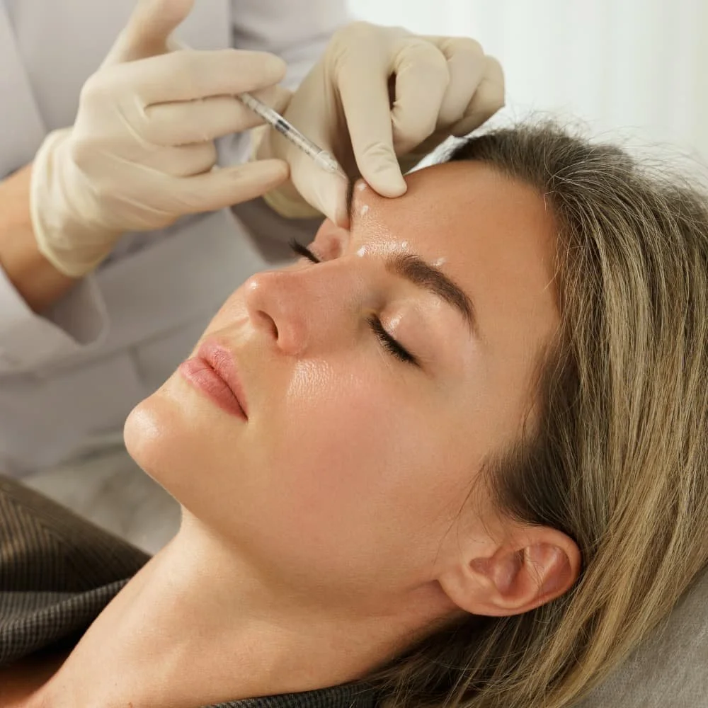A woman receives a cosmetic injection in her forehead from a medical professional wearing gloves.
