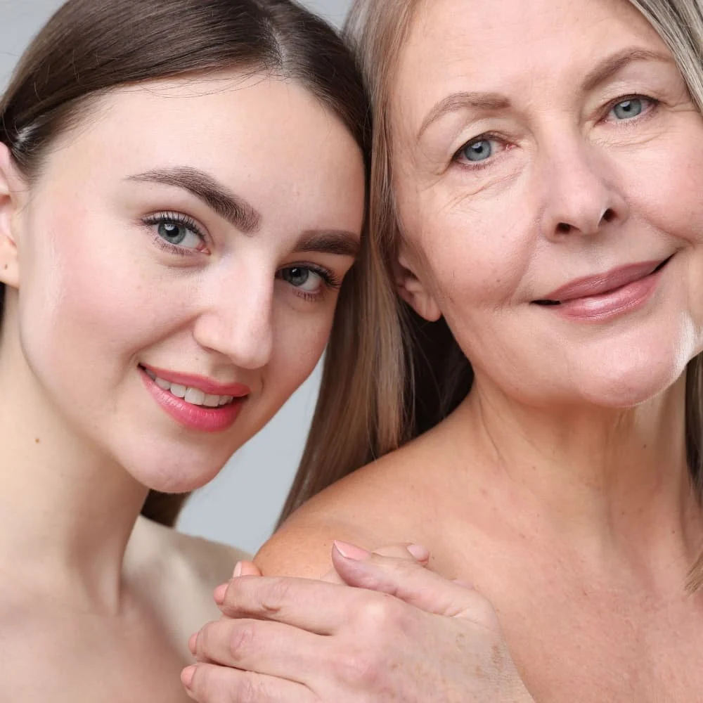 Close-up of a younger woman and an older woman smiling together, with the younger woman gently resting her hand on the older woman's shoulder.