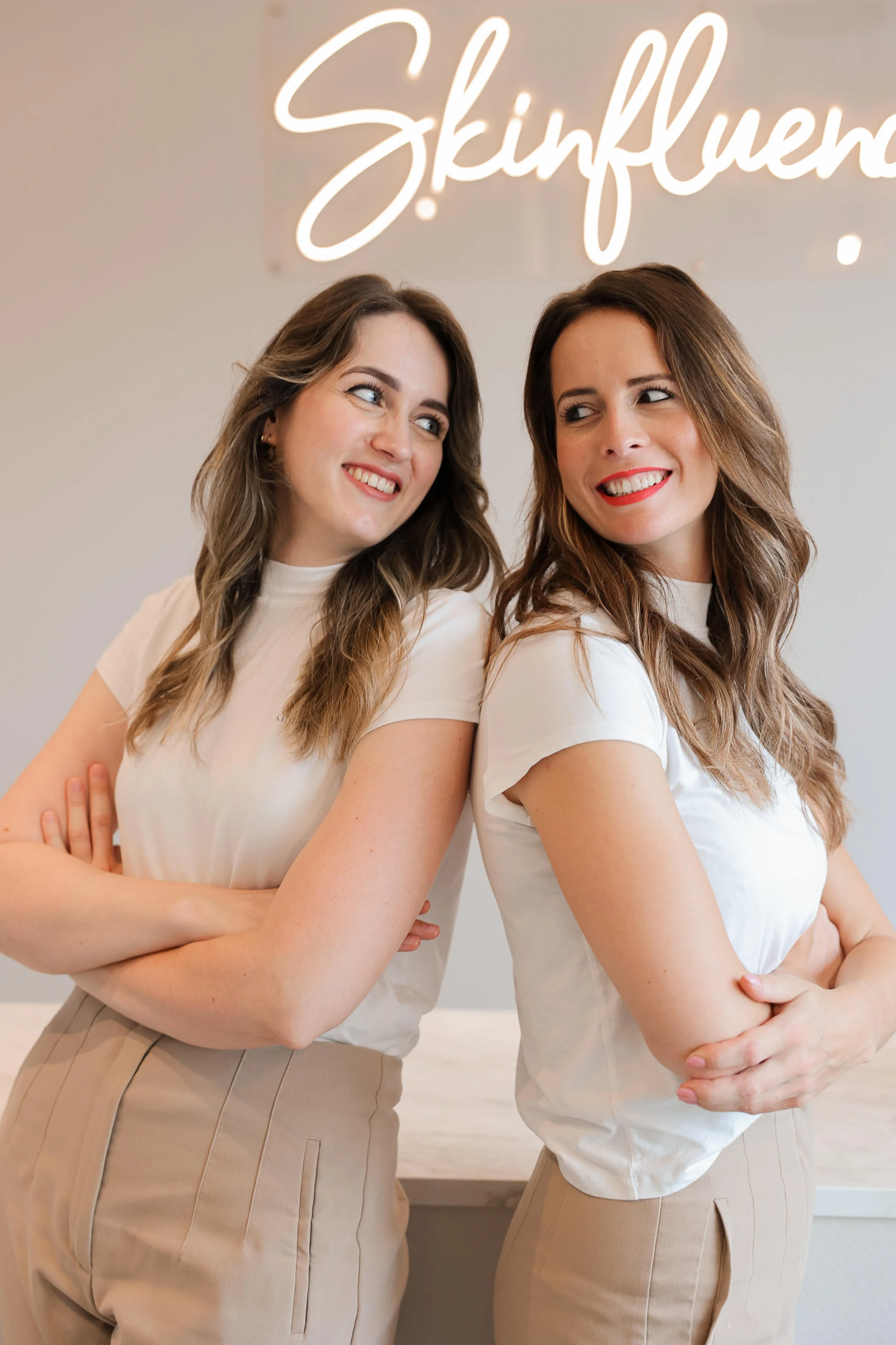 Twee vrouwen staan dicht bij elkaar met armen over elkaar, lachend en kijkend naar elkaar, voor een witte achtergrond met een neonlicht dat de naam 'Skinkluencers' uitstraalt.