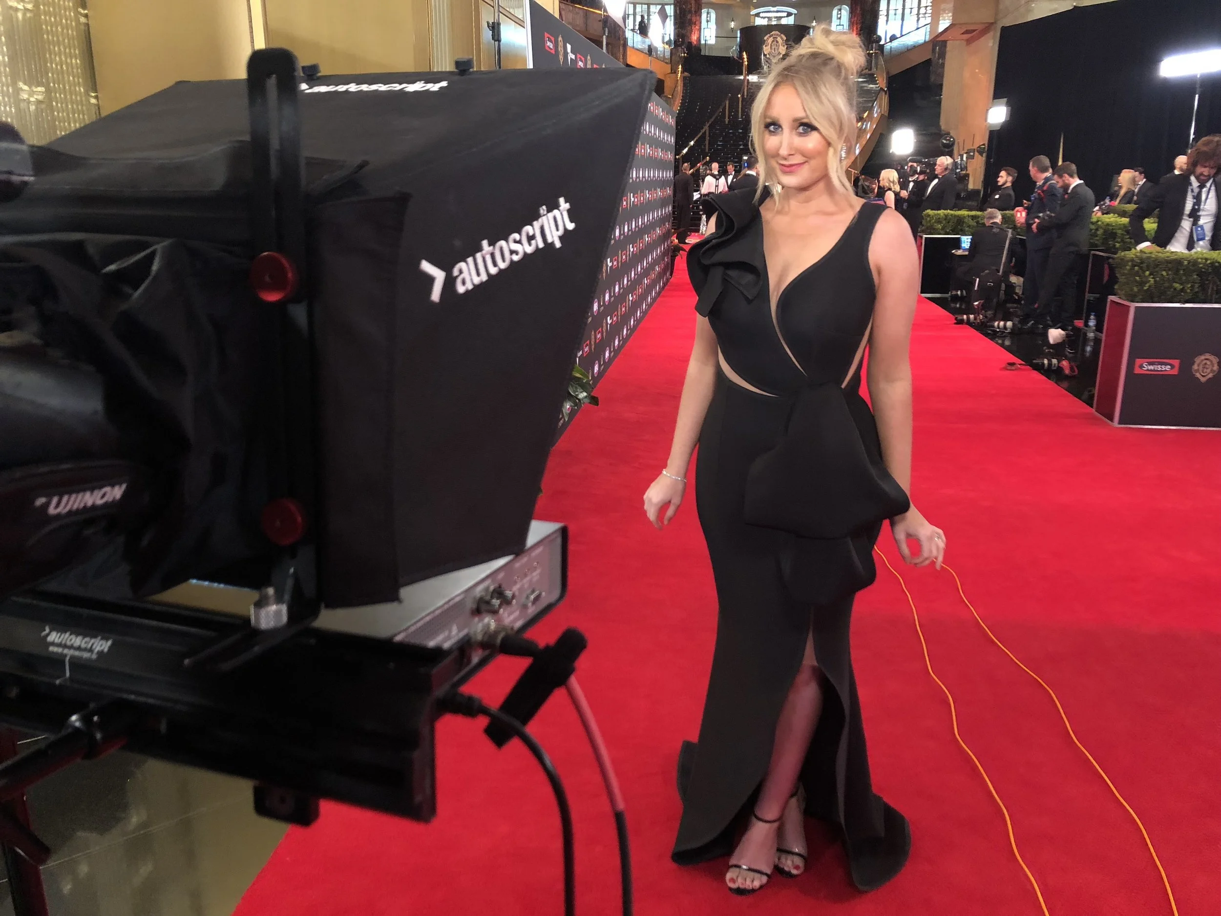 Jane Bunn on a red carpet at an event, wearing a black evening gown with a high slit, standing in front of a camera braodcasting 7NEWS at the Brownlow Awards, with a crowd and lighting setup in the background.