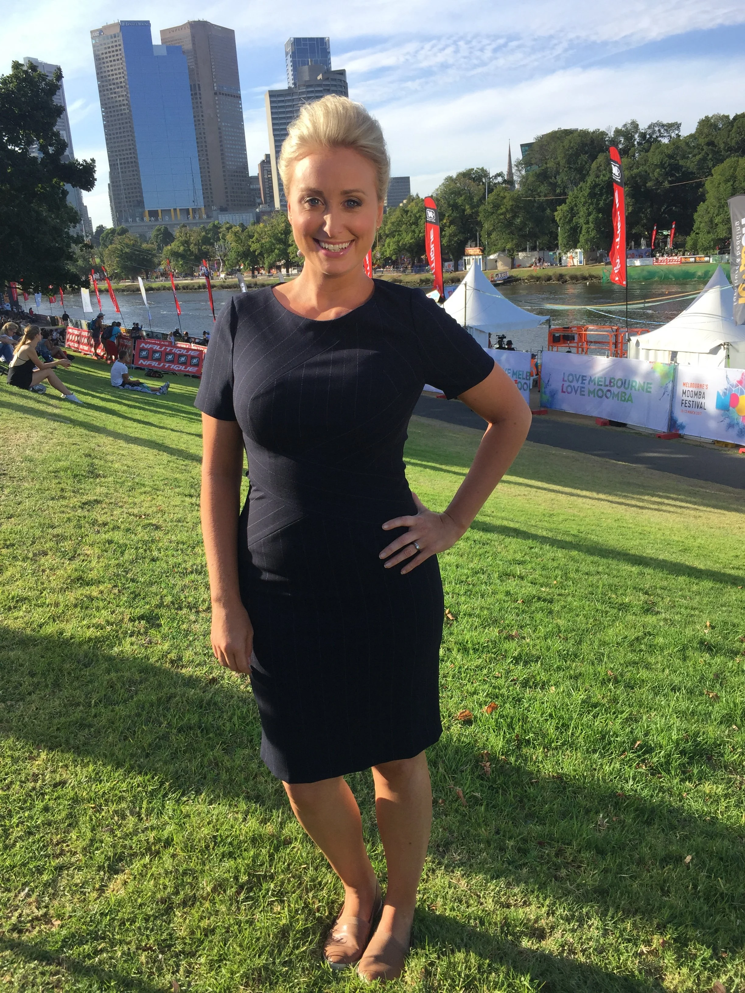 Jane Bunn in a black dress smiling and posing with one hand on her hip near a river, with tents, flags, and a city skyline in the background.