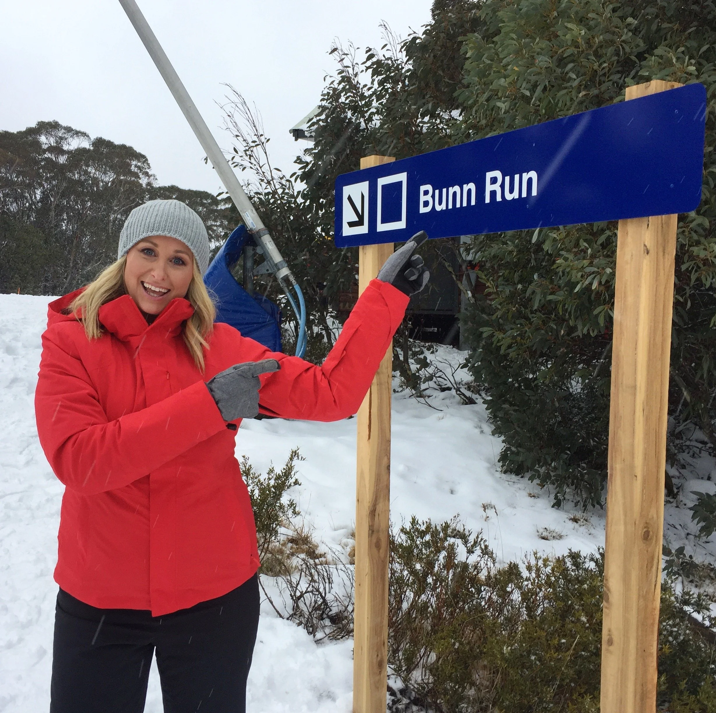 Jane Bunn in a red jacket and grey beanie pointing at a blue and white ski run sign that reads "Bunn Run" at Mt Buller.
