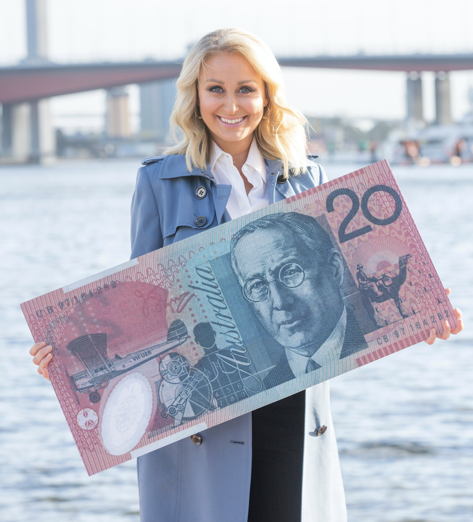 Jane Bunn in a blue coat holding an oversized Australian twenty-dollar bill for the RFDS near a body of water, with a bridge and city skyline in the background.