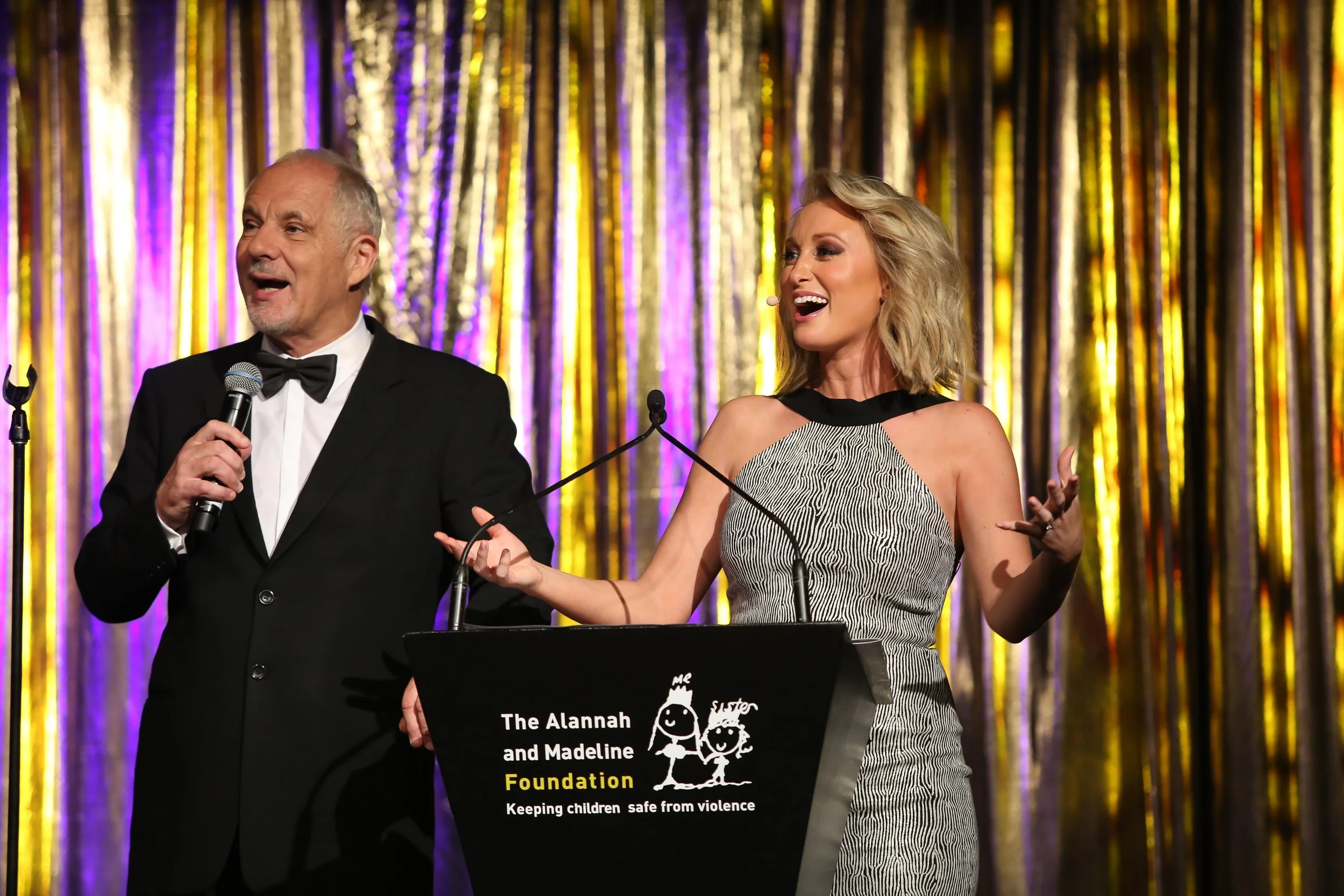 Jane Bunn speaking at a podium during an event with a golden curtain backdrop. The podium features a logo for The Alannah and Madeline Foundation, which promotes keeping children safe from violence.