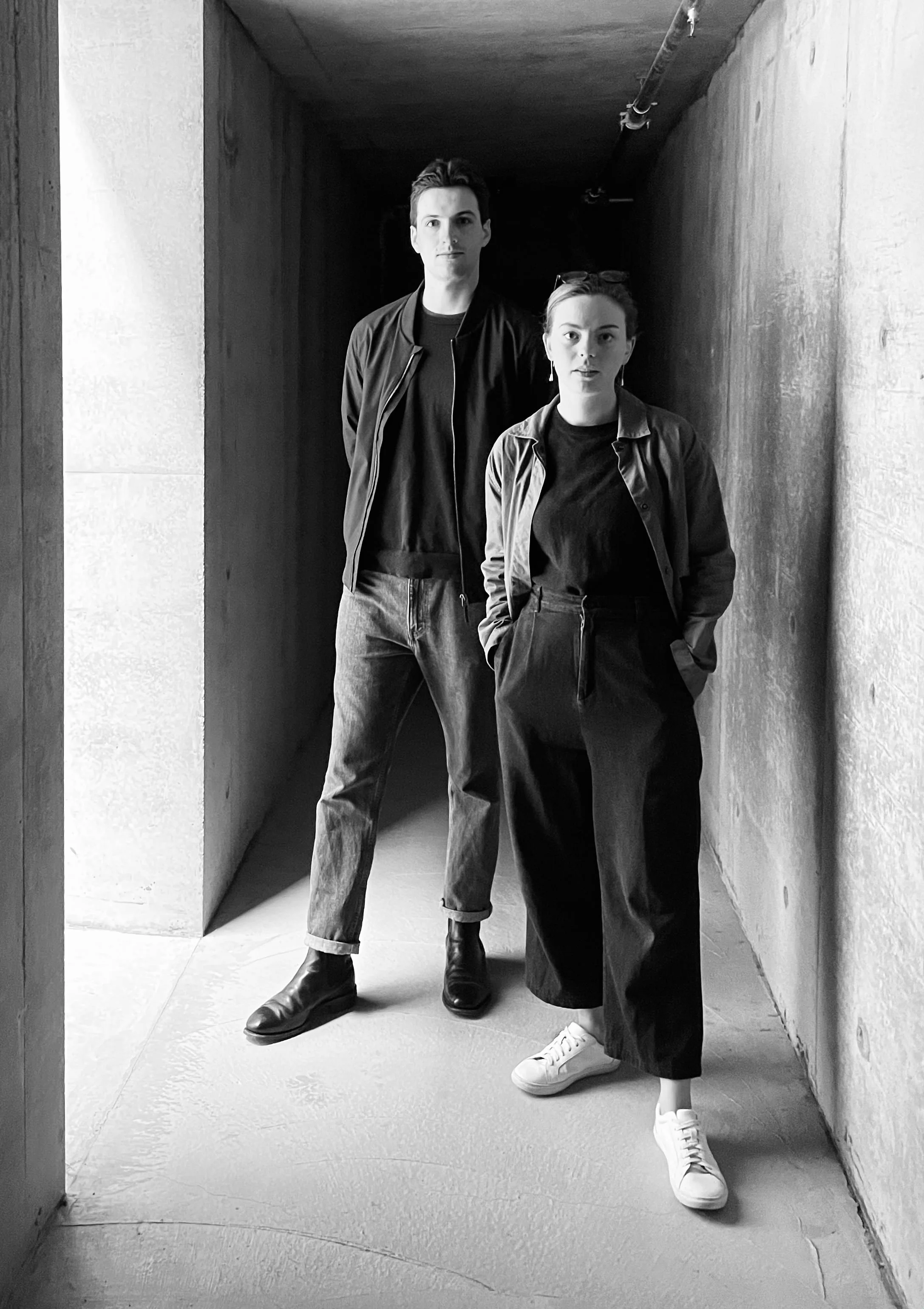 A black-and-white photo of a young man and woman standing in a narrow, dimly lit corridor. The man is tall with dark hair, wearing a jacket, t-shirt, jeans, and boots. The woman has dark hair in a bun, wearing a jacket, t-shirt, wide-legged pants, and white sneakers. Both are looking directly at the camera with neutral expressions.