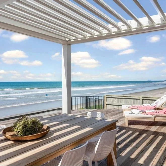 Beachside patio with wooden deck, outdoor dining table, white chairs, lounge chair with pink towel, view of ocean, blue sky with scattered clouds.