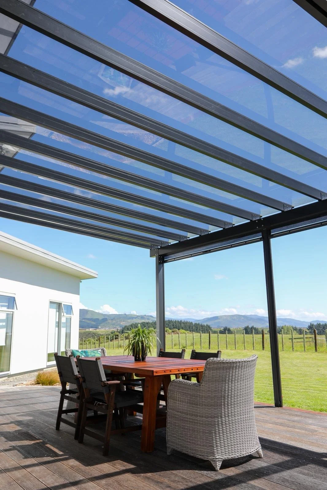 Outdoor patio with wooden table, wicker chair, and potted plant, shaded by a metal and glass pergola, overlooking a grassy field and mountains in the distance on a sunny day.