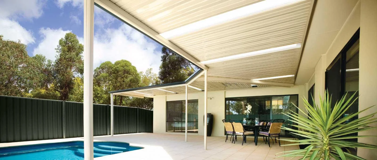 Empty outdoor patio with a swimming pool, covered by a cream-colored canopy, and outdoor dining table with six chairs. Green trees and blue sky in background.