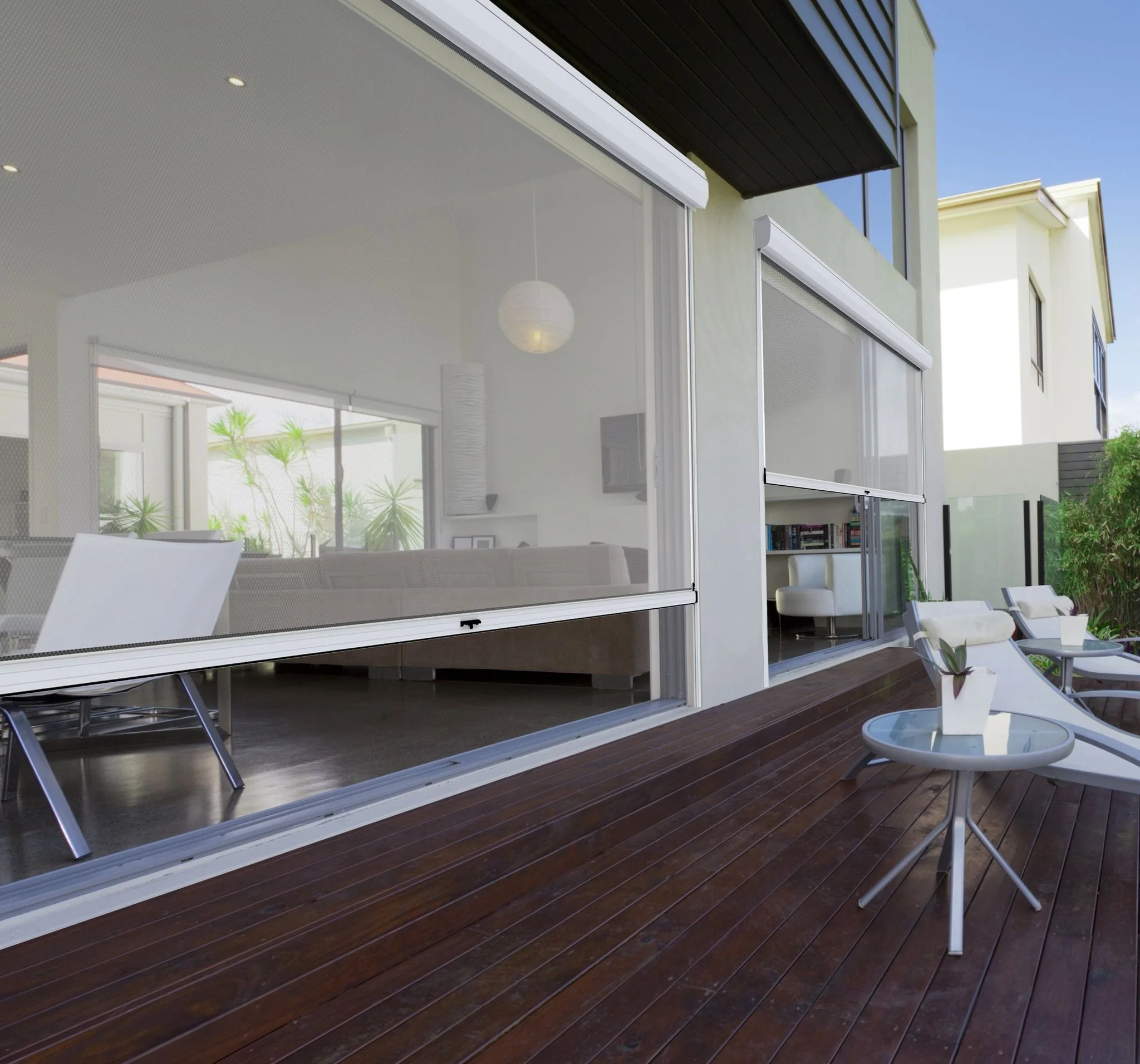 Modern house with sliding glass doors opening to a wooden deck with outdoor chairs and small side tables, backyard with green plants, and contemporary interior with white furniture and decor.