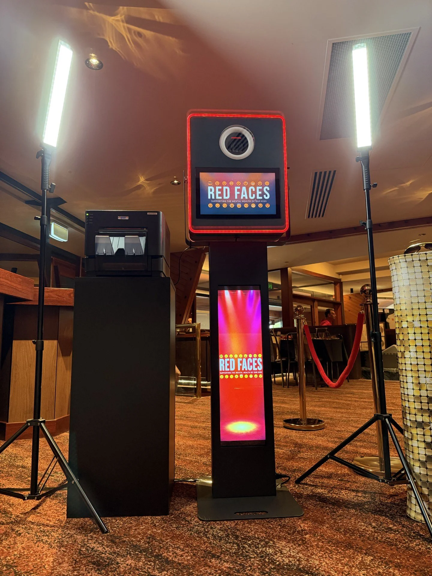 Photo of a digital display board with red border and text reading "Red Faces," supporting mental health, in a restaurant or event space, surrounded by professional lighting and velvet rope barriers.