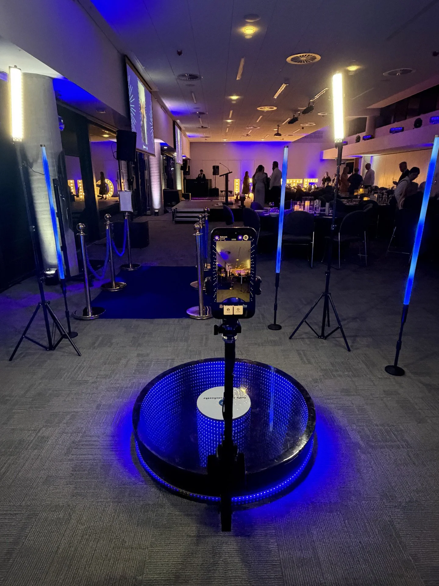 Empty event room with a DJ booth, tables, chairs, and blue lighting, set up for a celebration or party.