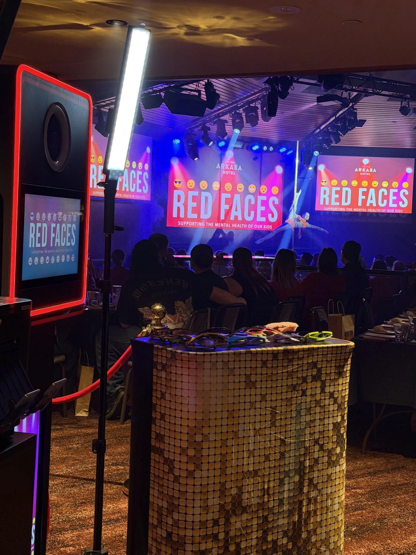 Stage with performer dancing under colorful lights, large screens displaying 'Red Faces' and supporting mental health of children, audience seated watching, event at Arkaba Hotel.