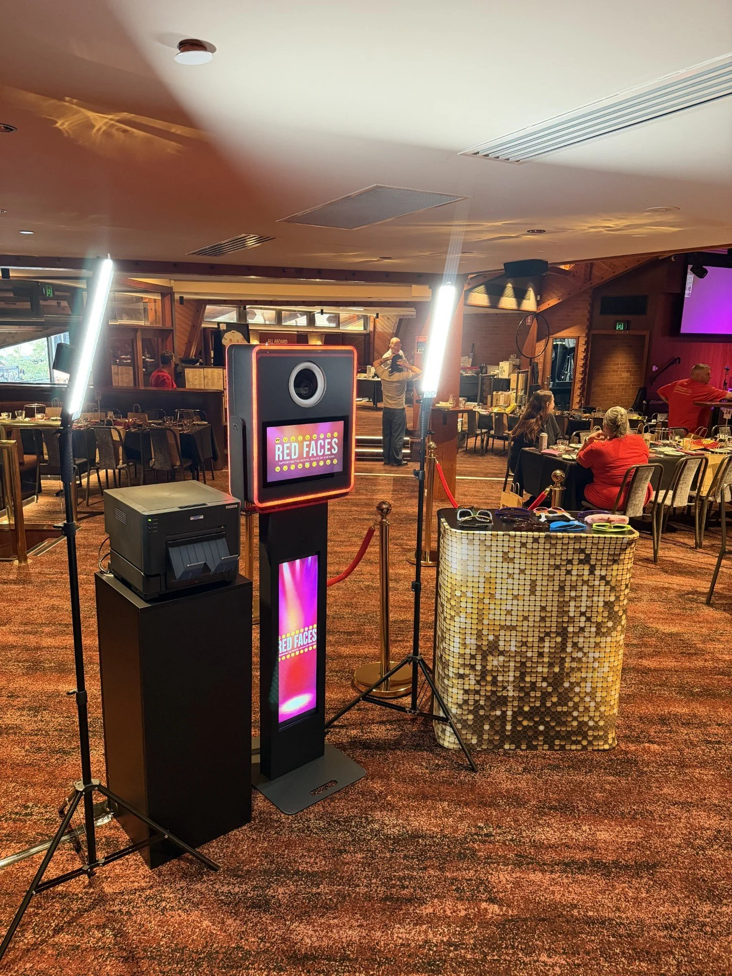 Indoor event space with tables, chairs, and lighting. A photo booth with a sign 'Red Faces' is set up in the foreground. Several people are seated and standing, some looking at their phones or talking. There's a stage area with a person speaking or s
