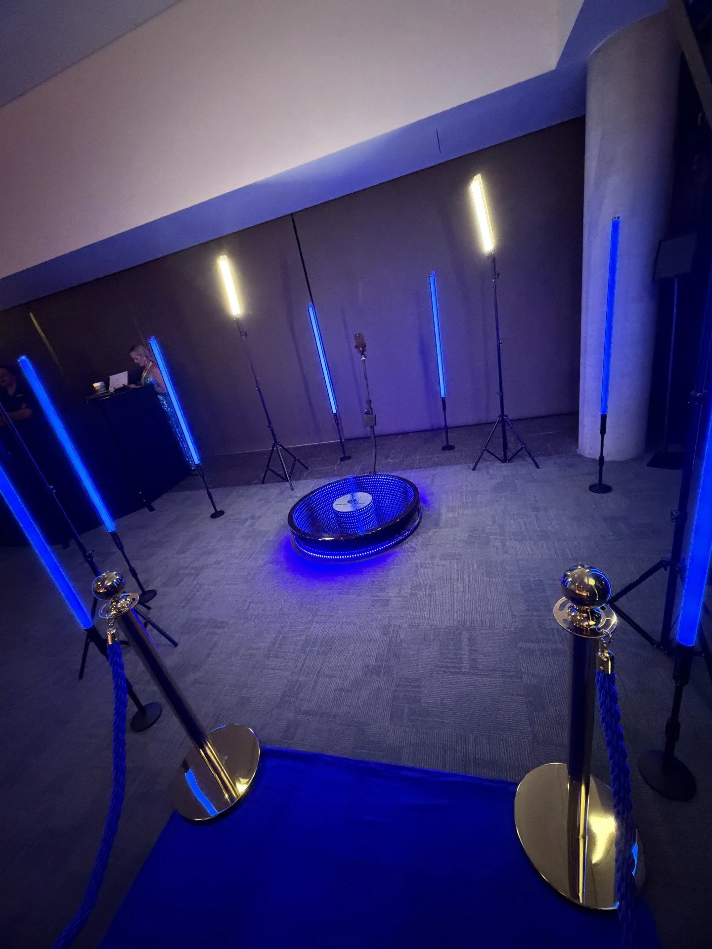 Room set up with blue and yellow lighting, a circular area with a low table, and stanchions with blue ropes surrounding it, possibly for a photo opportunity.