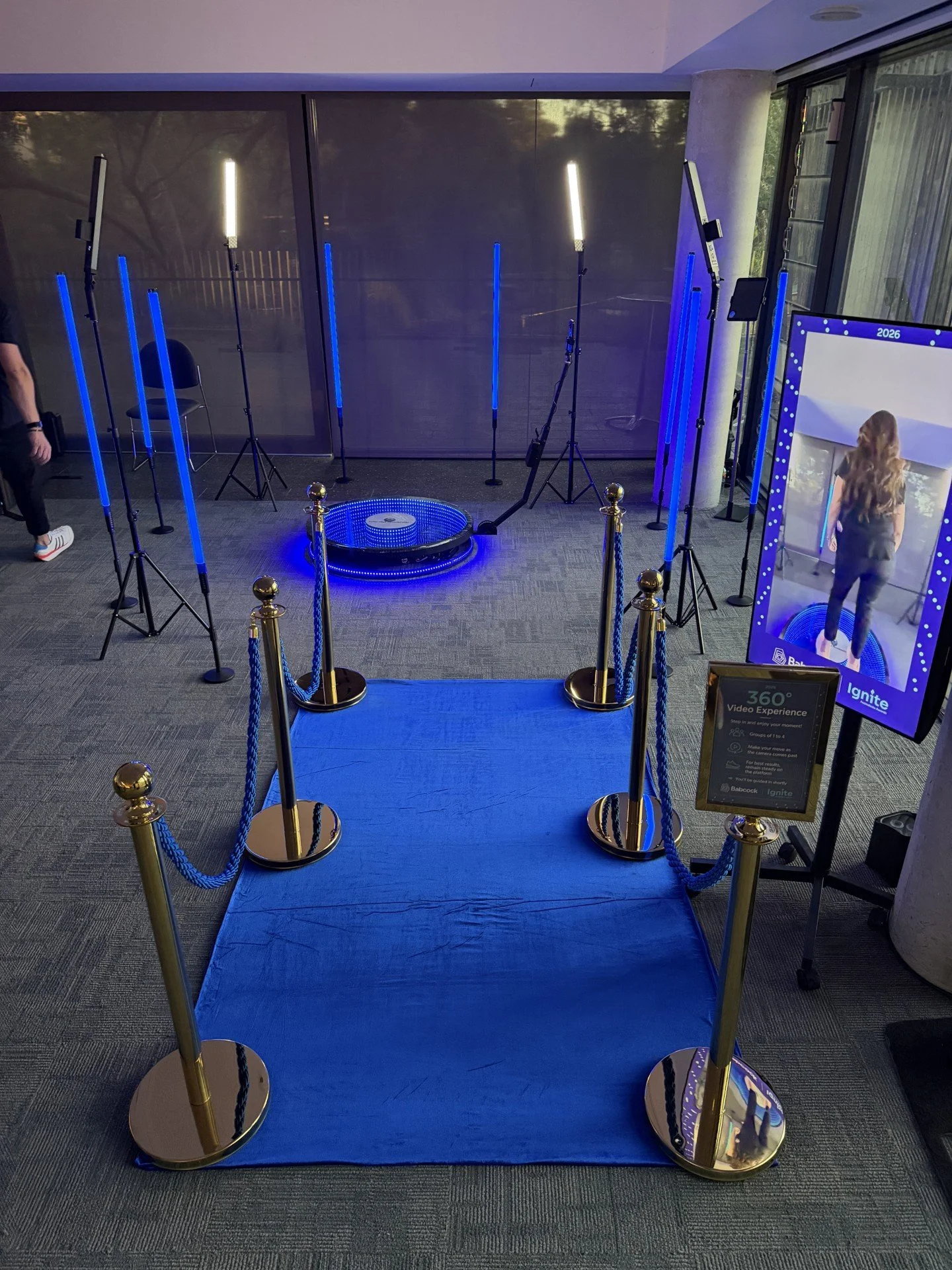 A blue-lit photo booth setup with a blue carpet and gold stanchions with blue ropes. There is a large mirror on the right, reflecting a person with wavy hair and text indicating it offers a 360-degree video experience. Several vertical LED light bars