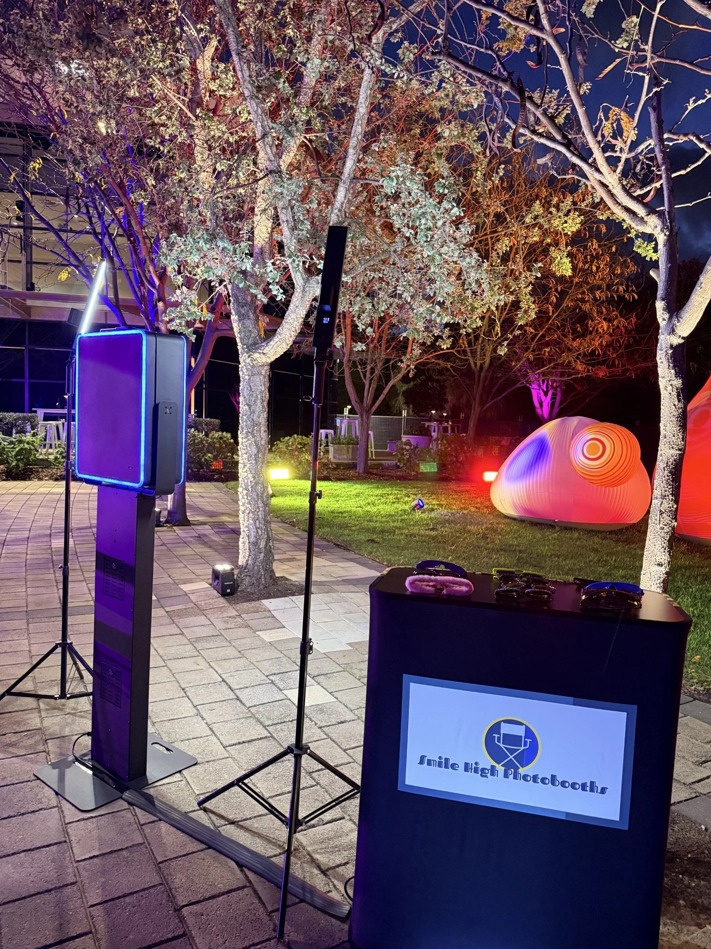 Nighttime outdoor scene with a photo booth setup, including a sign that reads "Jumble High Photobooths". There are trees with colored lights, a glowing spherical sculpture, and a lit-up area with some stools in the background.