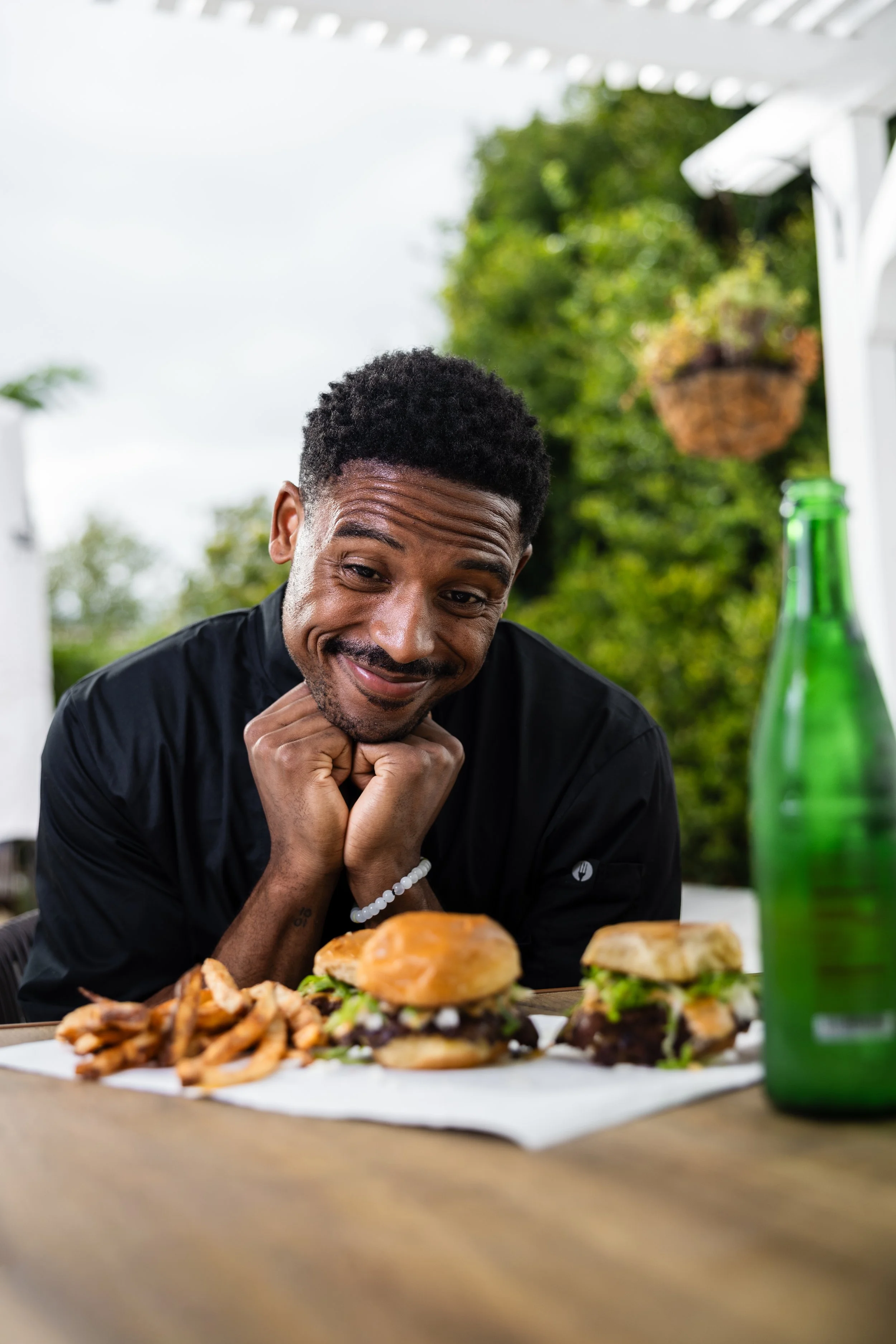 A man with short curly hair and a black shirt is smiling and leaning on a table with two burgers, a bunch of fries, and a green bottle. He is outdoors with green trees in the background.