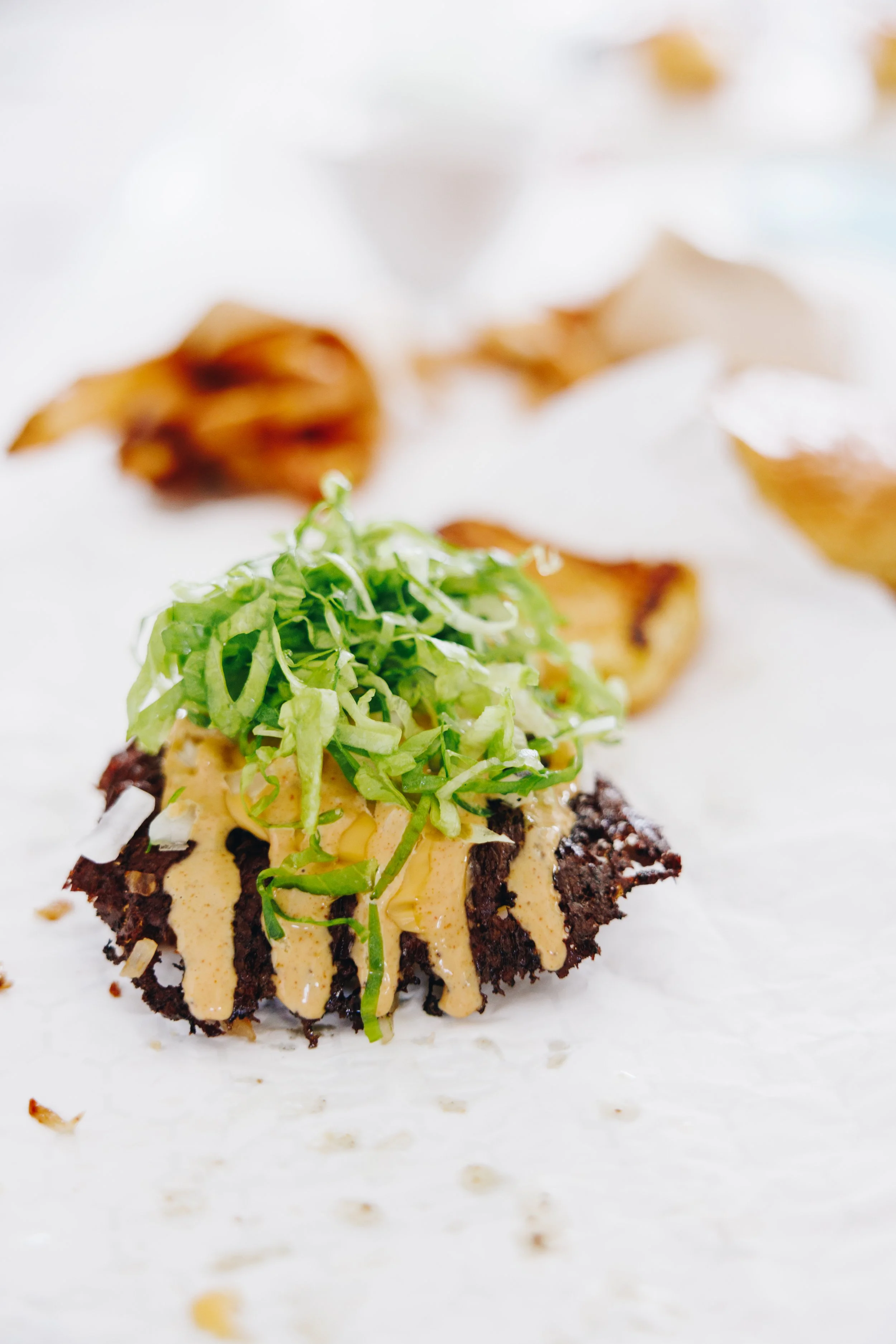 Close-up of a WOMO Burger with shredded lettuce, onions, cheese and a creamy sauce, with fries in the background on white paper.