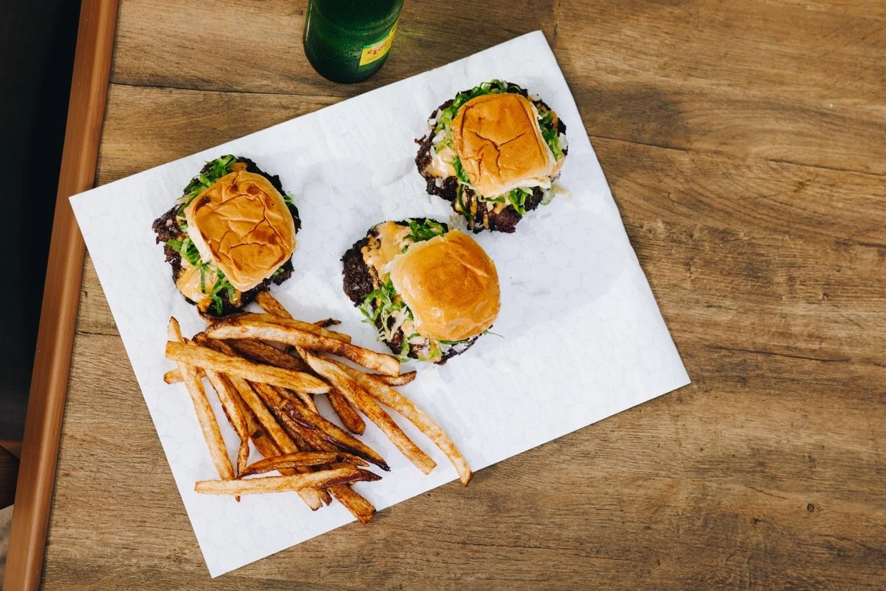 Three sliders with buns, lettuce, and meat, served with a side of French fries on paper, and a bottle and cup in the background on a wooden table.