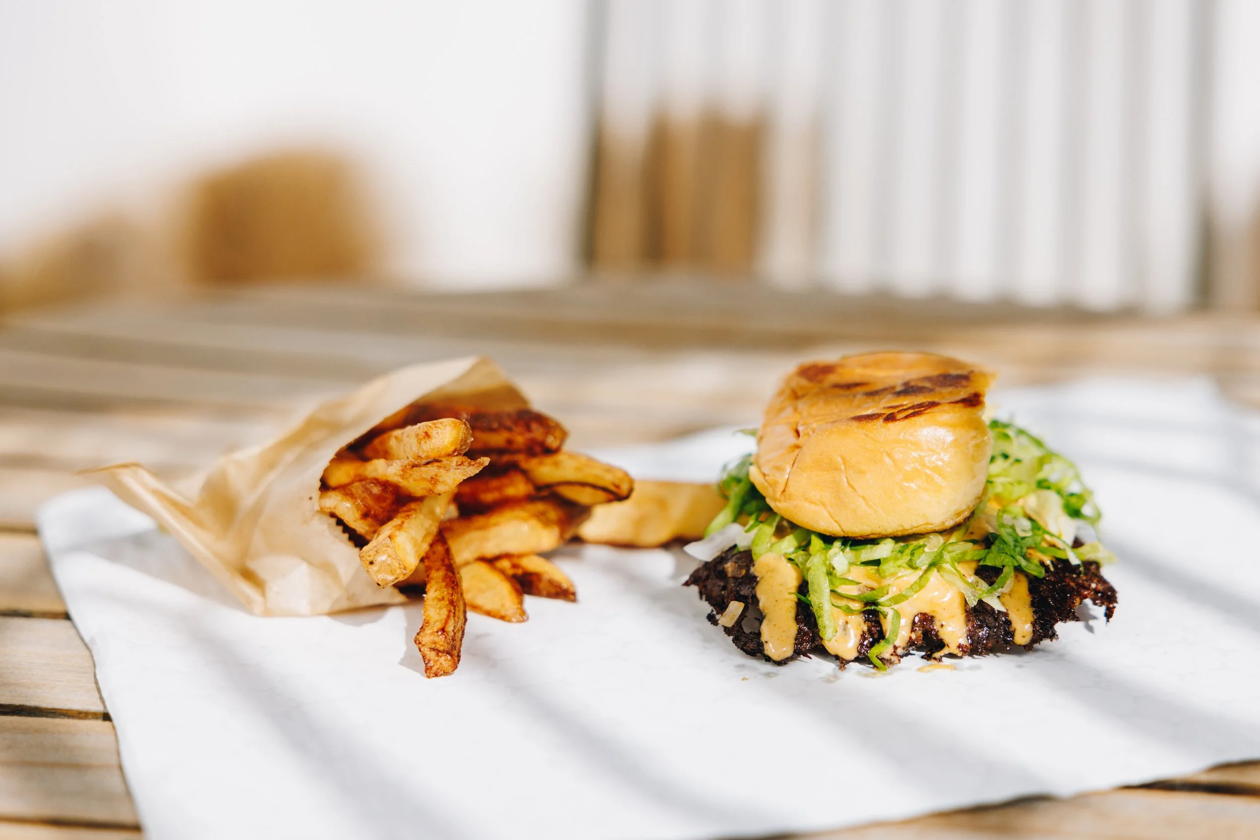 A burger with lettuce, cheese, and a grilled patty topped with a bun, served with a side of French fries on white paper on a wooden table.