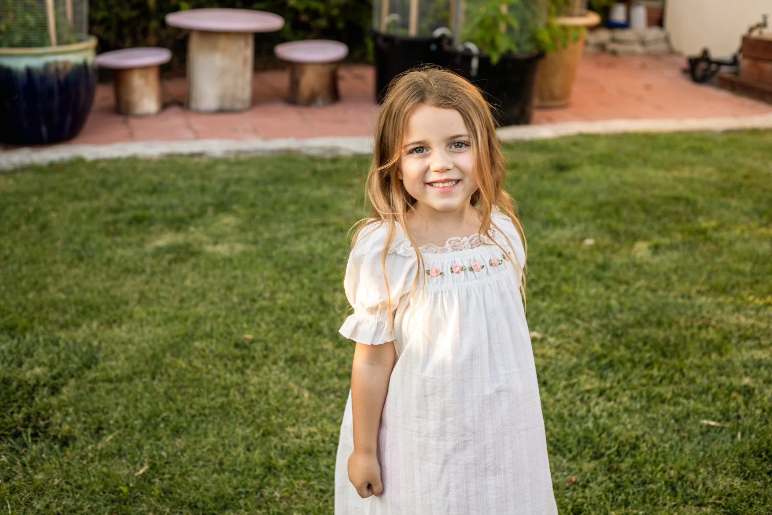 A young girl with long, wavy hair and a bright smile standing on a grass lawn, wearing a white dress with floral embroidery near the neckline, in a backyard setting with potted plants and outdoor furniture in the background.