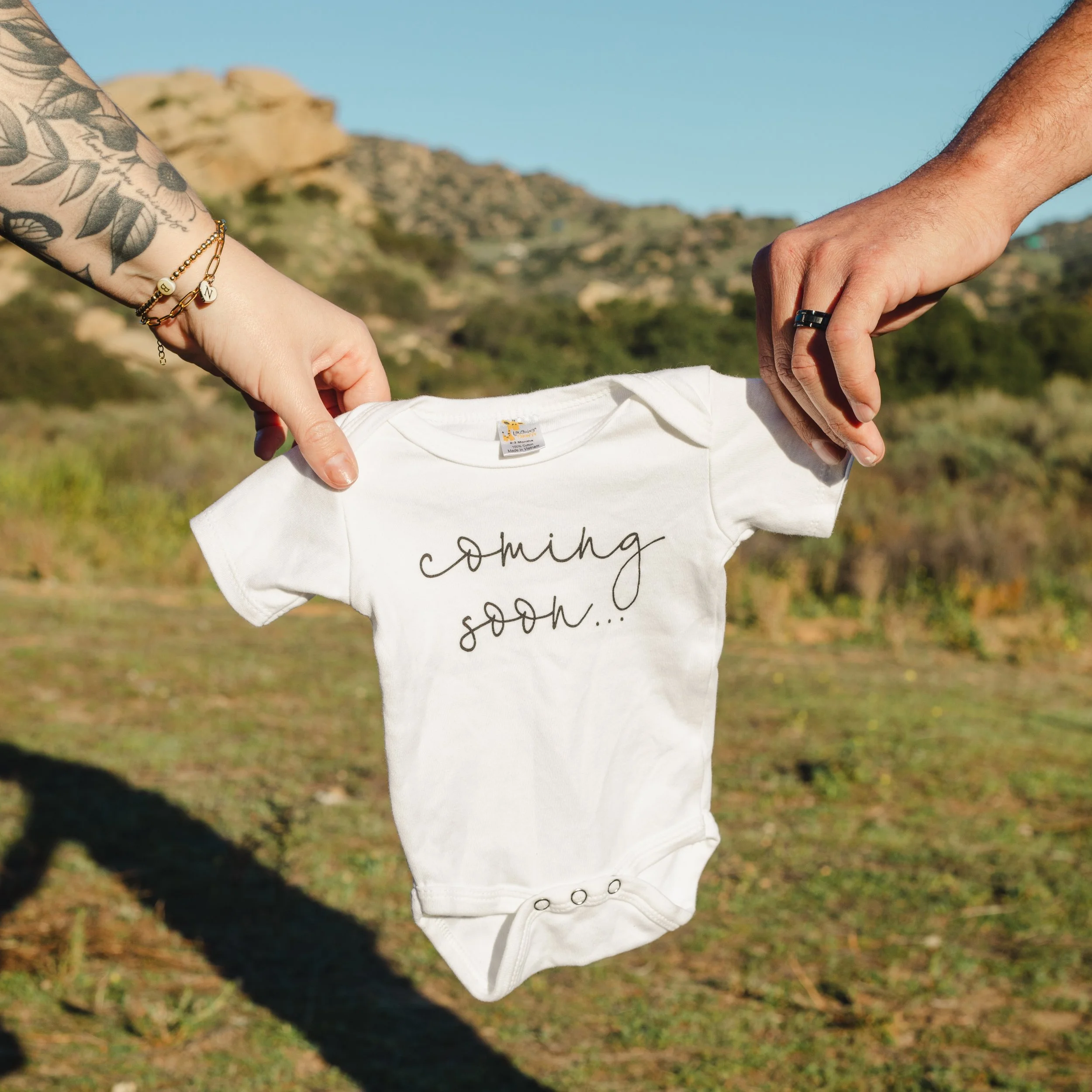 Two people hold up a white baby onesie with the text 'coming soon...' written on it, outdoors with hills and trees in the background.
