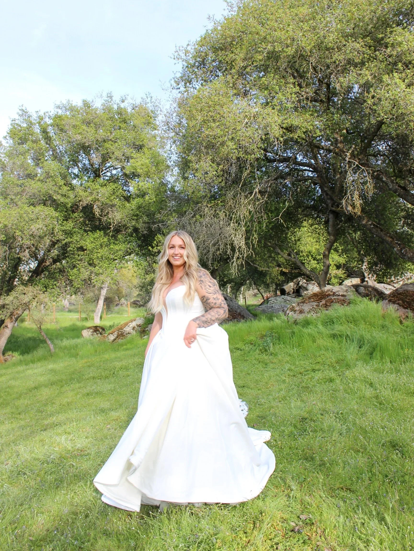 A smiling woman in a white wedding dress standing on green grass with trees and rocks in the background.