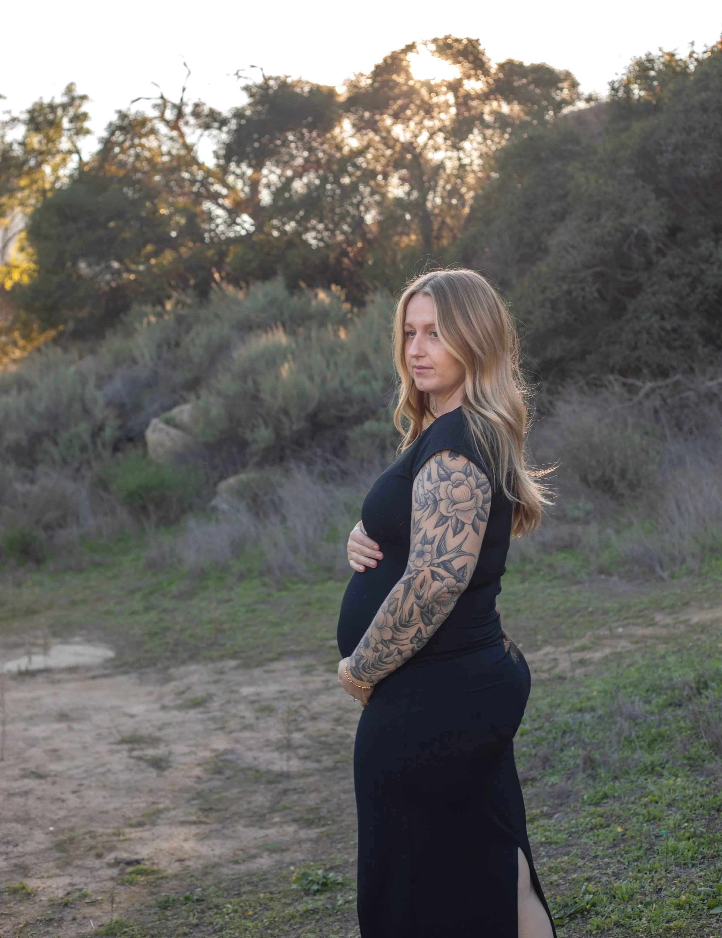 A pregnant woman with long blonde hair and a floral tattoo on her left arm stands outdoors in a natural setting during sunset, wearing a black dress with a side slit.