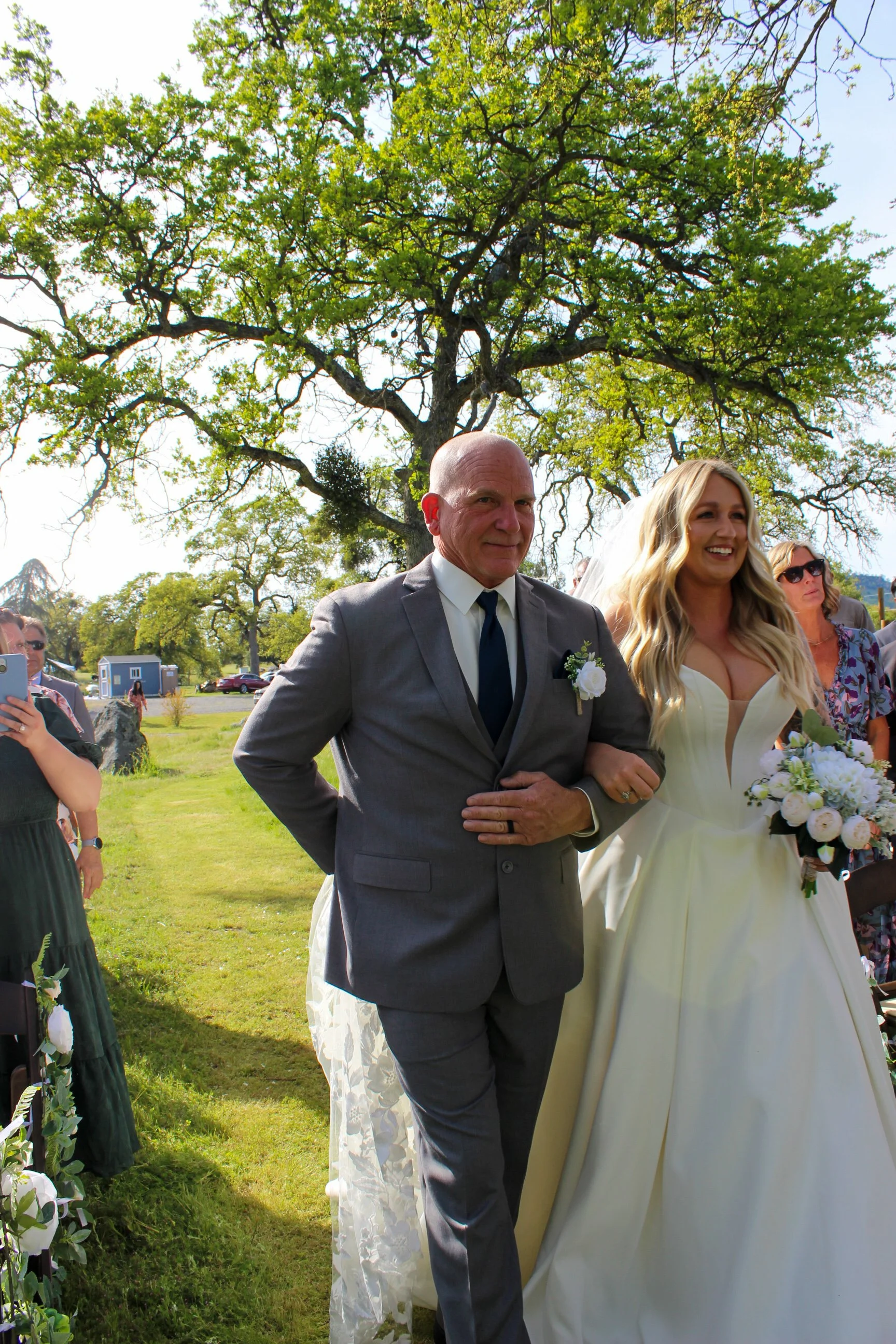 A bride walking down the aisle with a man, likely her father, outdoors on a sunny day, with guests watching and taking pictures in the background.