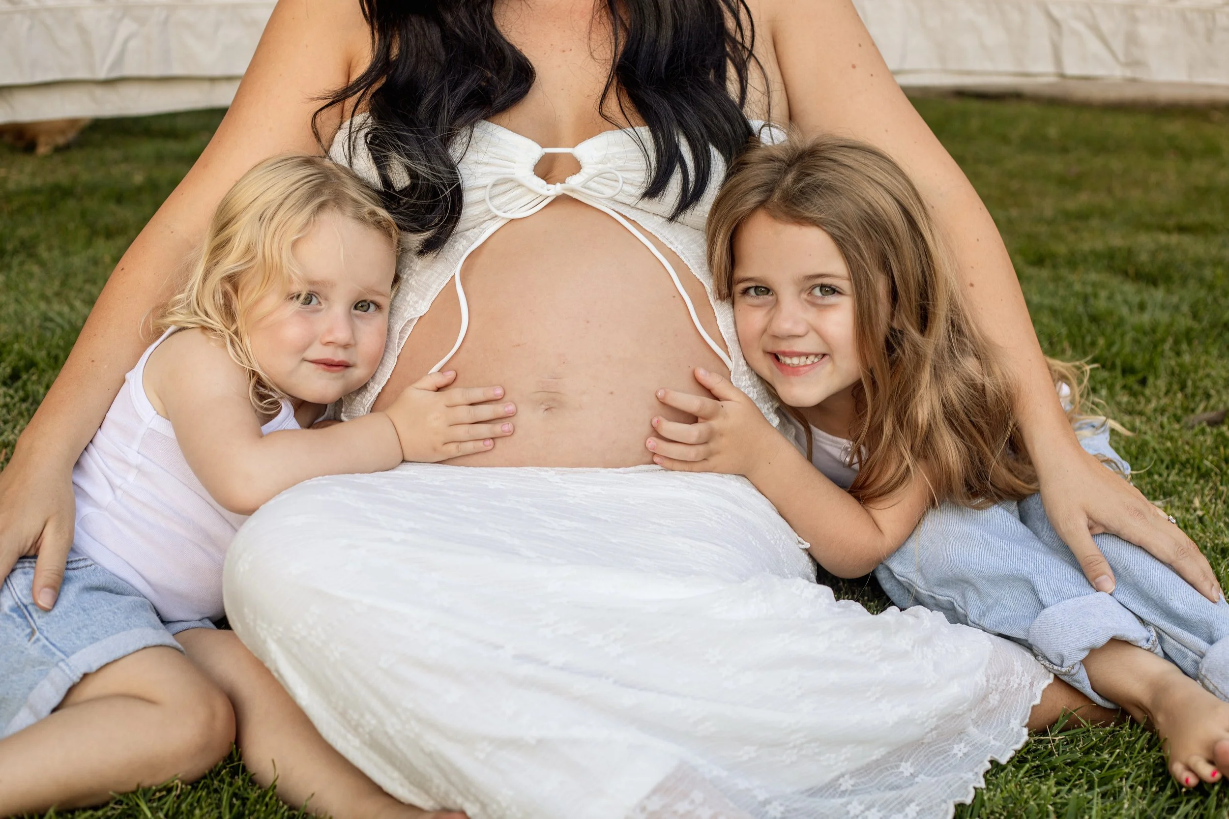 A pregnant woman sitting on grass outdoors with two young girls hugging her belly, smiling and looking at the camera.