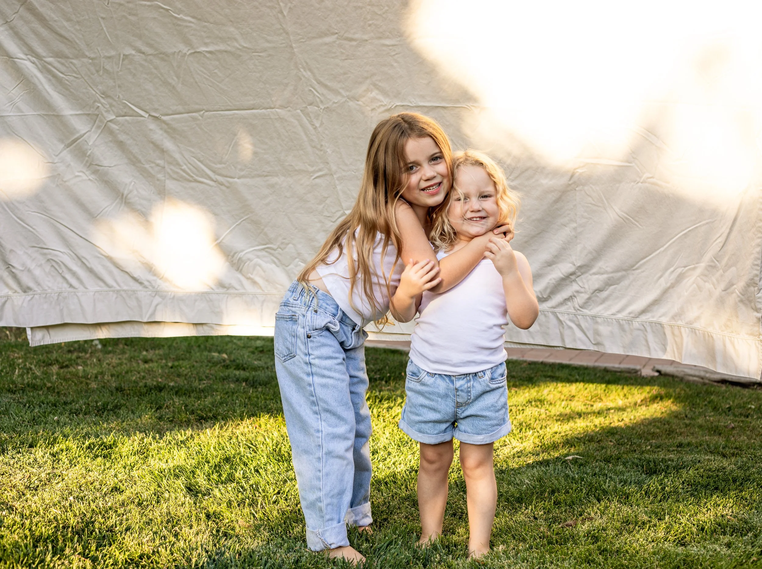 Two young girls hugging each other and smiling on a grassy lawn with a cream-colored backdrop behind them.